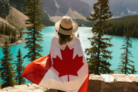 A person draped in a Canadian flag stands on a stone ledge looking out at a turquoise glacial lake surrounded by pine trees and mountains The persons back is to the camera and they are wearing a hat Bathed in warm sunlight this breathtaking moment could be part of your dream Canada tour package