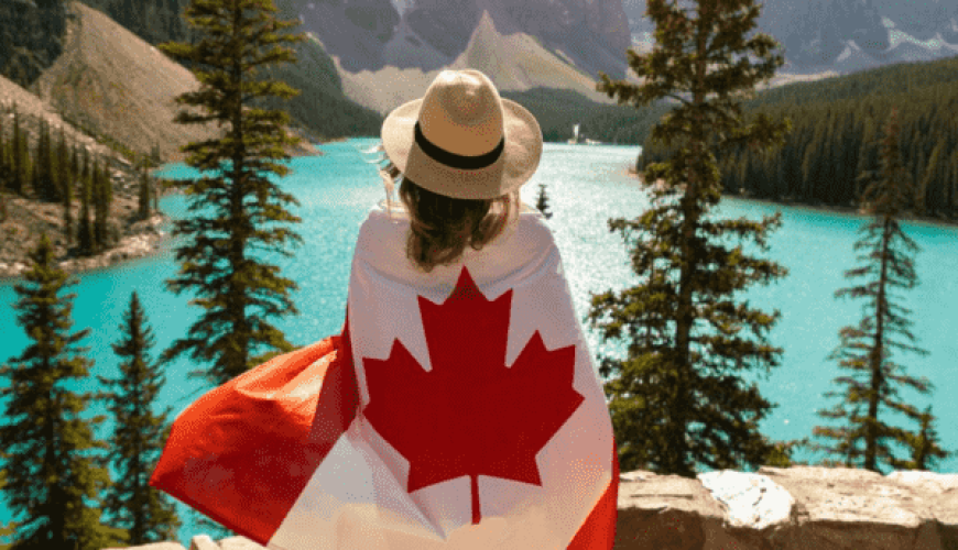 A person draped in a Canadian flag stands on a stone ledge, looking out at a turquoise glacial lake surrounded by pine trees and mountains. The person's back is to the camera, and they are wearing a hat. Bathed in warm sunlight, this breathtaking moment could be part of your dream Canada tour package.